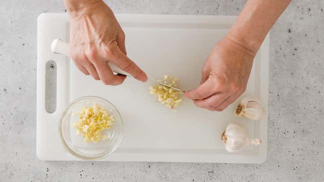 Hands cutting garlic on a plastic cutting board.